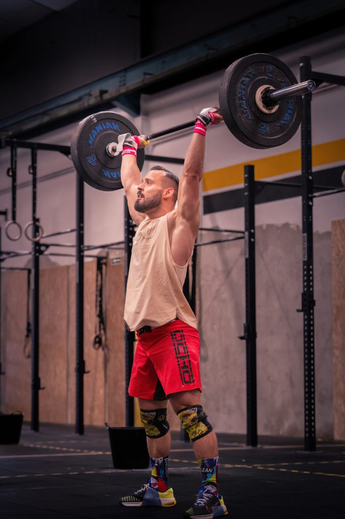 Un hombre vestido de deportivo levanta una barra por encima de la cabeza en un gimnasio, de pie sobre un suelo negro con equipos de ejercicio en el fondo.