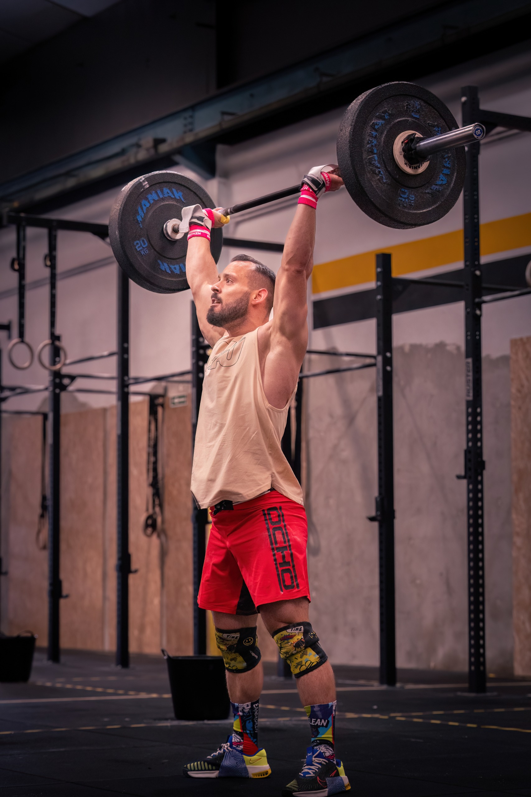 Un hombre vestido de deportivo levanta una barra por encima de la cabeza en un gimnasio, de pie sobre un suelo negro con equipos de ejercicio en el fondo.