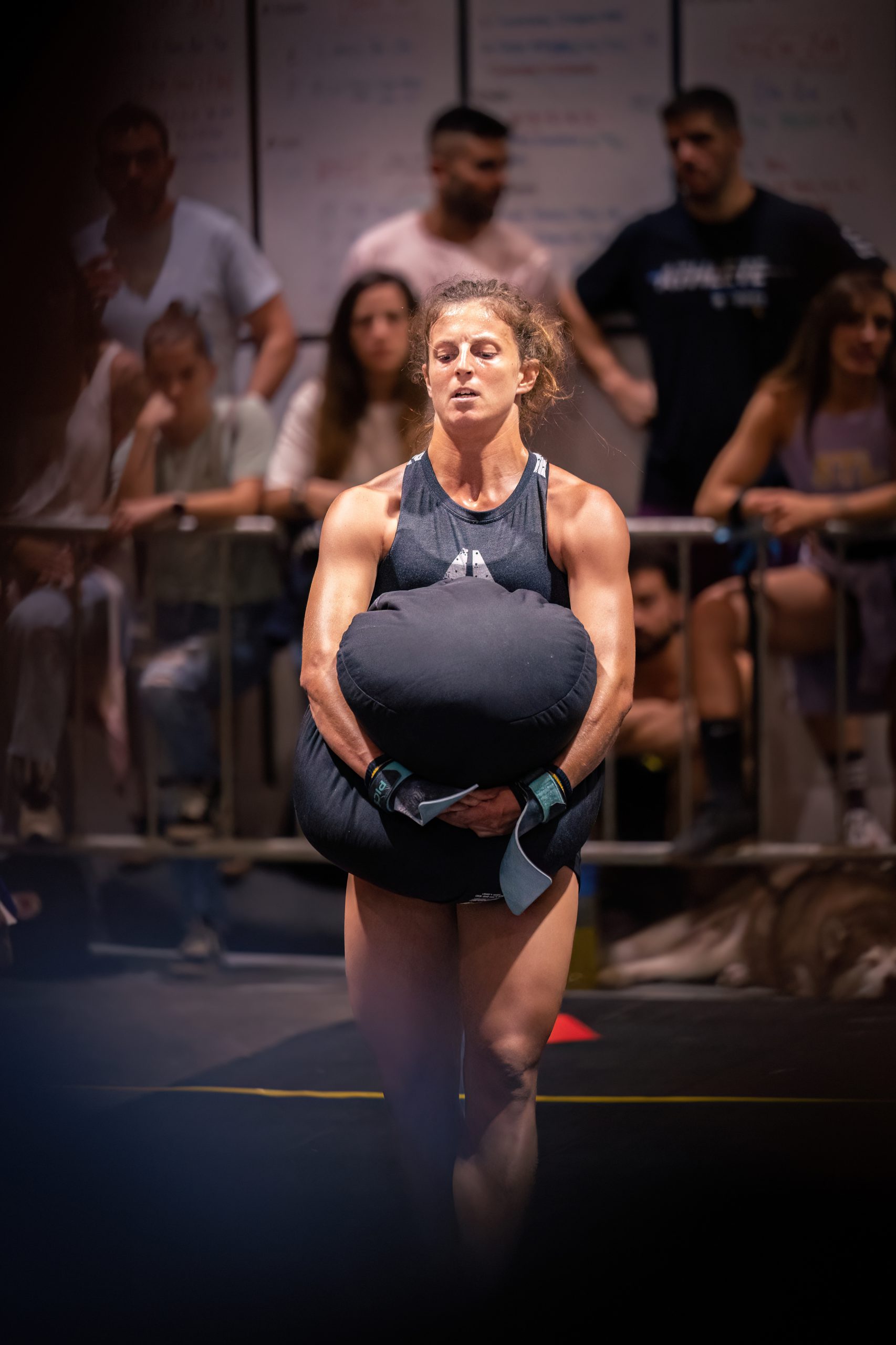 Una mujer vestida con ropa deportiva lleva un pesado saco de arena durante una competición de fitness, mientras los espectadores la observan desde el fondo.