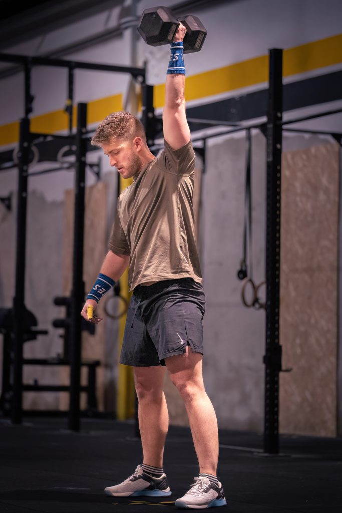 Un hombre con ropa deportiva se encuentra en un gimnasio, sosteniendo una mancuerna sobre su cabeza con un brazo.