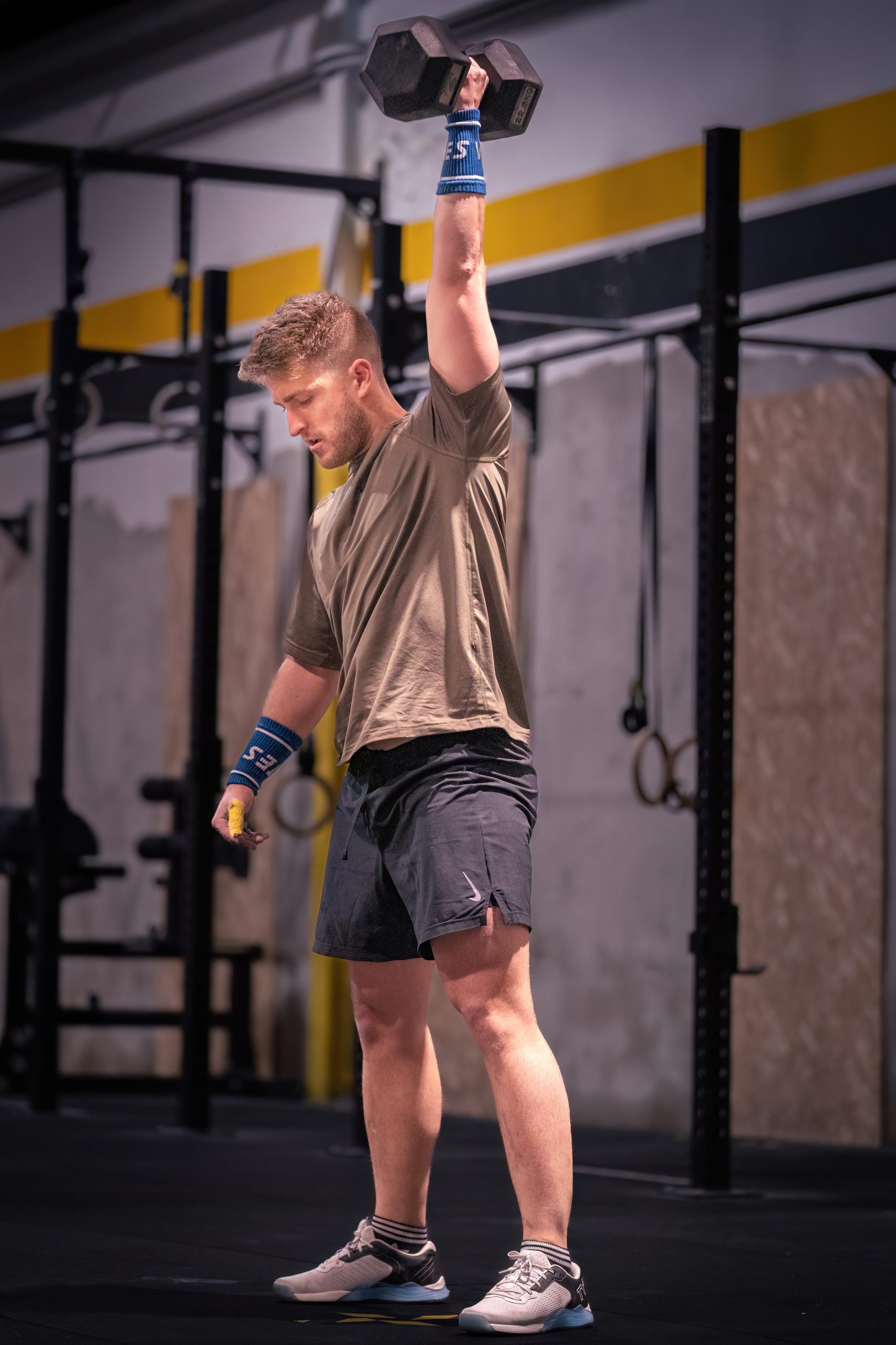 Un hombre con ropa deportiva se encuentra en un gimnasio, sosteniendo una mancuerna sobre su cabeza con un brazo.